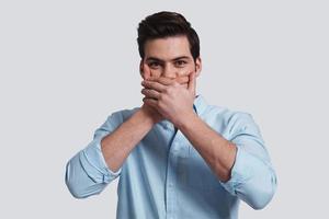 Amazing news  Handsome young man covering mouth with hands and looking at camera while standing grey background photo