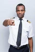piloto serio. piloto africano serio en uniforme apuntando la cámara mientras está de pie contra el fondo gris foto