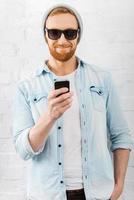 Quick text. Happy young bearded man holding mobile phone and smiling while standing against brick wall photo