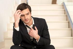 Sick and tired. Depressed young man in formalwear holding head in hand and taking off his necktie while sitting on staircase photo