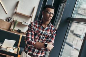 Thinking about... Serious young man keeping arms crossed and looking away while standing near the window photo