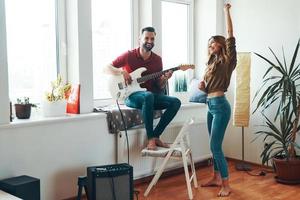 Pareja joven juguetona con ropa informal uniéndose y sonriendo mientras toca la guitarra foto