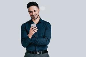 buenas noticias de un amigo. un joven guapo sosteniendo un teléfono inteligente y mirándolo con una sonrisa mientras se enfrenta a un fondo gris foto