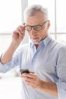 Reading new business messages. Serious grey hair senior man in shirt holding mobile phone and looking at it while standing in front of the window photo