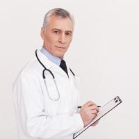 Prescribing the proper medicine. Senior grey hair doctor in uniform writing something in clipboard and looking at camera while standing isolated on white photo