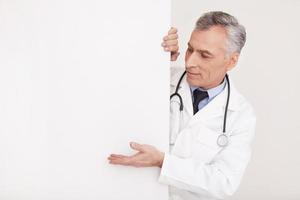 Doctors advice. Senior grey hair doctor in uniform looking out of copy space and pointing it while isolated on white photo