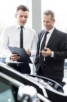 Some paperwork before buying a car. Handsome young classic car salesman talking to a customer and pointing a note pad photo