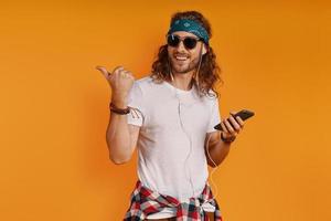 Handsome young man listening music and pointing copy space while standing against yellow background photo