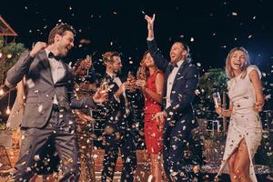 Group of beautiful people in formalwear dancing and smiling while confetti flying all around photo