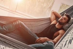 Simply resting. Top view of handsome young man in casual clothing relaxing while lying in the hammock indoors photo