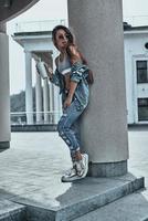 Beauty and style. Full length of attractive young woman in jeans wear looking away and holding a disposable cup while leaning on the architectural column outdoors photo