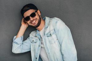 Loving his style.Cheerful young man adjusting his baseball cap and looking at camerawith smile while standing against grey background photo