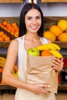 The bag full of health. Beautiful young woman in apron holding paper shopping bag with fruits and smiling while standing in grocery store with variety of fruits in the background photo