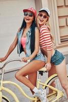 Summertime is fun time. Cheerful young woman sitting on bicycle while her female friend standing behind her photo