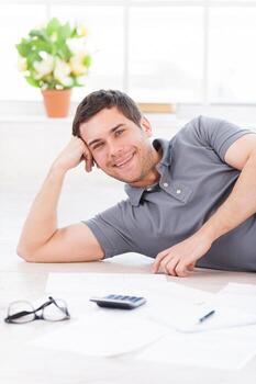 I used to work at home. Cheerful young man holding hand on chin and smiling while lyingon the floor with calculator and documents on itI used to work at home. Cheerful young man holding hand on chin and smiling while lyingon the floor with calculator and photo