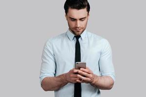 Quick chat with business partner. Serious young man in formalwear typing text message using his smart phone while standing against grey background photo