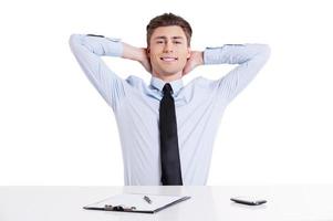 Feeling calm and confident. Handsome young man in shirt and tie sitting at the table and holding hands behind head while isolated on white photo