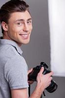 fotógrafo alegre. joven con camisa de polo sosteniendo una cámara digital y mirando por encima del hombro mientras estaba de pie en el estudio con equipo de iluminación en el fondo foto