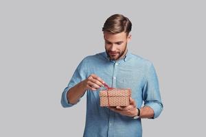 Interesting...  Handsome young man tying gift box and smiling while standing against grey background photo