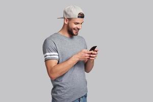 Receiving nice messages.  Handsome young man holding smart phone and looking at it while standing against grey background photo