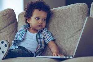 Digital age kid. Little African baby boy using his laptop while sitting on the couch at home photo