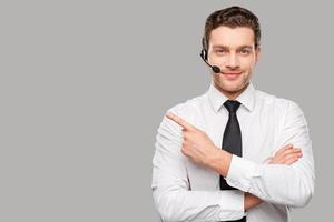 Operator pointing copy space. Handsome young man in formalwear and headset looking at camera and pointing away while standing against grey background photo