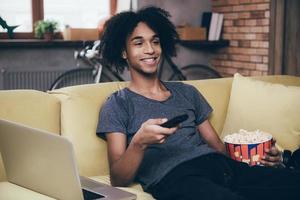 Surfing the channels. Cheerful young African man watching TV and holding remote control while sitting with bucket of popcorn on the couch at home photo