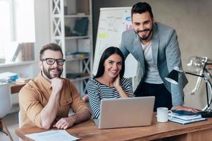 Successful team. Three confident business people in smart casual wear looking at camera and smiling while sitting at the desk in office photo