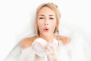 belleza tomando baño. mujer joven juguetona acostada en la bañera llena de espuma jabonosa y soplando un beso de aire foto