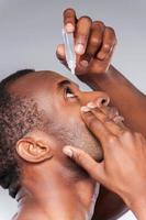 goteando sus ojos con gotas para los ojos. vista lateral del joven africano aplicando gotas para los ojos mientras está de pie contra el fondo gris foto