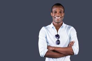 encantador guapo. un apuesto joven negro con camisa azul que mantiene los brazos cruzados y sonríe a la cámara mientras se enfrenta a un fondo gris foto