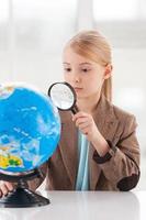 examinando el globo. niña concentrada en ropa formal examinando el globo con una lupa mientras se sienta en la mesa foto