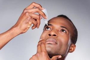 hombre goteando ojo. joven africano aplicando gotas para los ojos mientras se enfrenta a un fondo gris foto