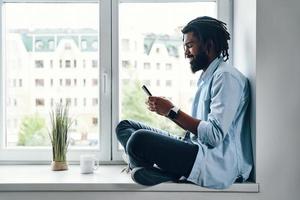 Happy young African man using smart phone and smiling while sitting on the window sill indoors photo