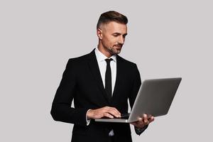 Modern technologies. Handsome young man in full suit using laptop and smiling while standing against grey background photo