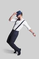 Perfect in every way. Full length of handsome young man in suspenders adjusting his hat and making a face while dancing against grey background photo