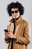 Message from friend.Cheerful young African man in sunglasses looking at camera with smile and holding smartphone while standing against grey background photo