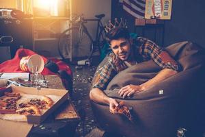 rey de la fiesta. Un joven apuesto con una corona de plástico tirado en una bolsa de frijoles con una rebanada de pizza en la mano en una habitación desordenada después de la fiesta foto