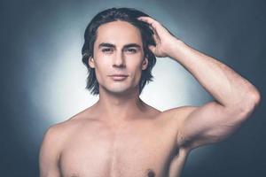 Looking just perfect. Portrait of young shirtless man looking at camera and adjusting his hairstyle while standing against grey background photo