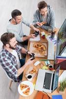 tiempo de juego. vista superior de tres jóvenes jugando juegos de computadora y comiendo pizza mientras están sentados en el escritorio foto