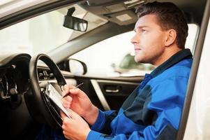 Man examining car. Confident young man examining car and writing something in clipboard while sitting in a car at workshop photo