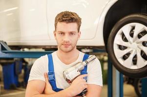 Confident mechanic. Confident young man holding a wrench and looking at camera while standing in workshop with car in the background photo