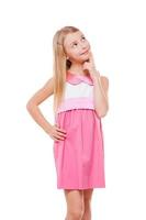 Thoughtful child. Cheerful little girl in pink dress holding hand on chin and looking away while isolated on white photo