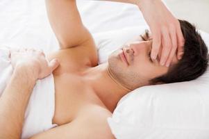 Insomnia. Handsome young shirtless man holding hand in hair and keeping eyes closed while lying in bed photo