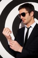 Confident in himself. Handsome young man in formalwear and sunglasses adjusting his shirt cuffs while standing against black and white background photo