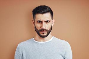 Frowning young man in casual wear looking at camera while standing against brown background photo