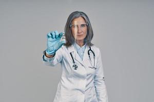 Mature beautiful female doctor in face shield holding a bottle with vaccine photo