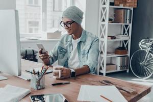 Receiving nice messages. Handsome young man holding a cup and looking at smart phone with smile while sitting at his working place in creative office photo
