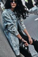 Enjoying carefree time outdoors. Attractive young woman in denim jacket looking at camera while leaning on the building outdoors photo