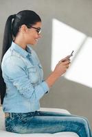 Great message from friend. Side view of beautiful young cheerful woman in glasses using her smart phone with smile while sitting on the couch against grey background photo